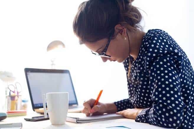 Woman wearing glasses and polka dot blouse sitting at desk and writing in notebook next to coffee mug and laptop computer.