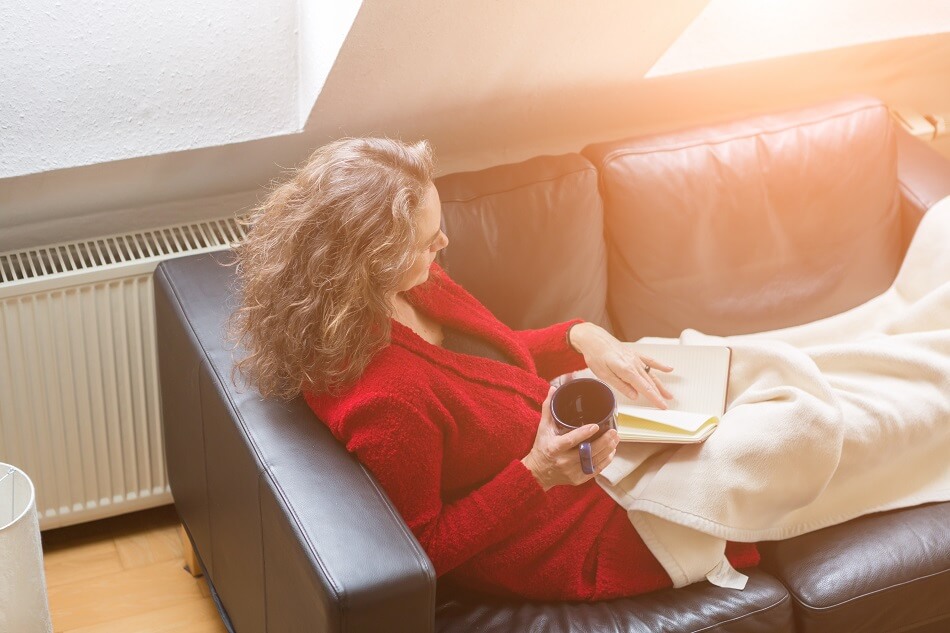 brunette woman wearing red sweater holding mug sitting on leather couch with blanket and journal while feeling lazy