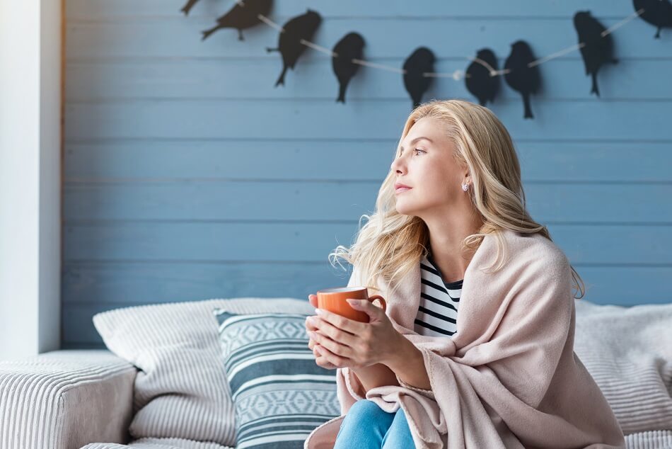 blonde woman holding orange mug sitting on couch looking into distance while considering what to do when feeling lazy