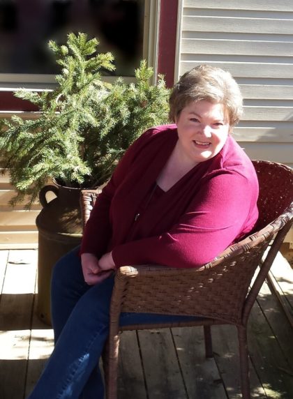 woman with short blond hair wearing jeans and red sweater sitting in wicker chair on porch against greenery and tan siding background