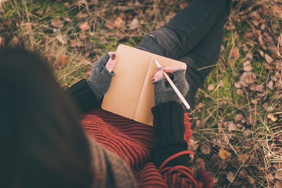 woman in red and black sweater and black jeans sitting on autumn grass and leaves while writing in journal