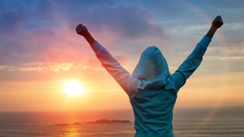 woman in hoodie with arms raised standing on beach at sunset