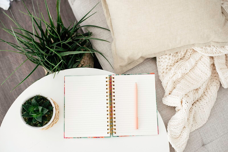 cozy couch with pillow and blanket next to plants and white table with open journal ready to use for february daily writing prompts