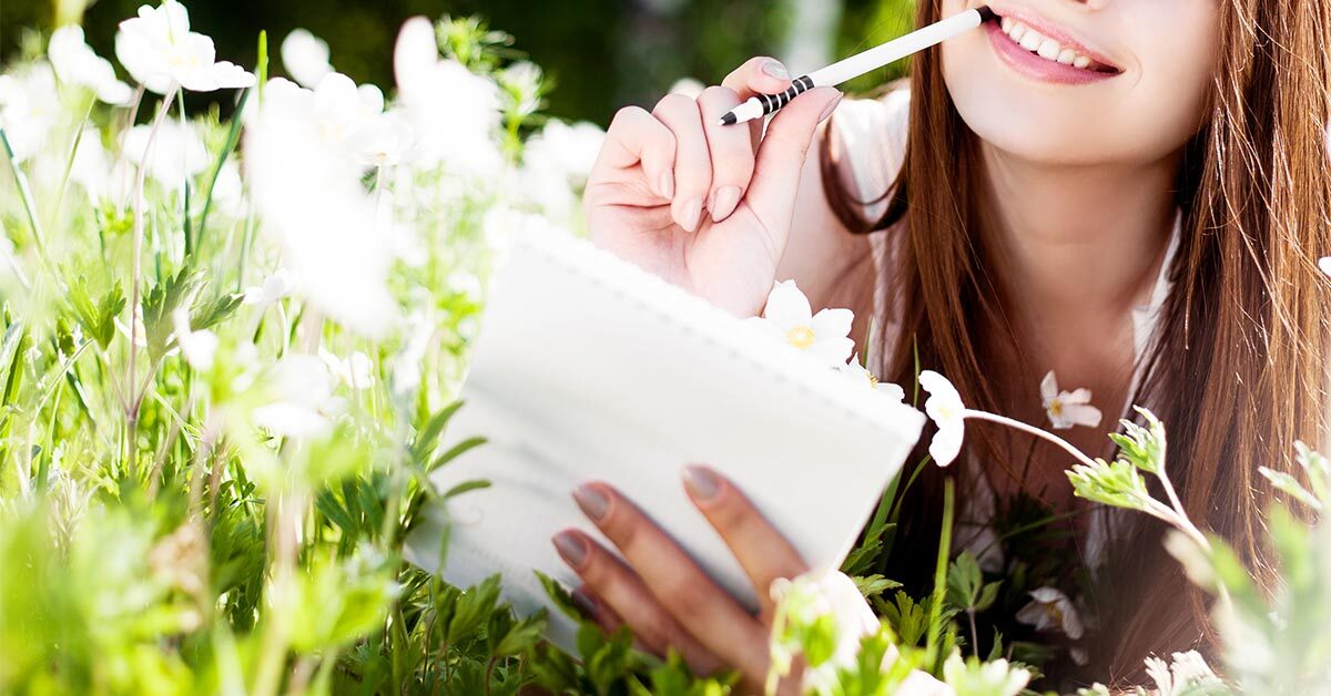 young woman lying in wildflower field on sunny day while getting ready to journal in notebook