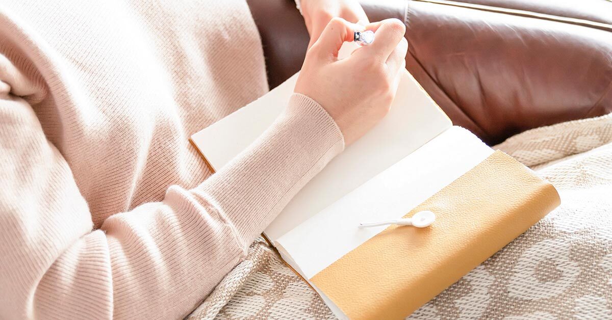 woman in pink sweater with blanket sitting on brown leather couch while writing in journal