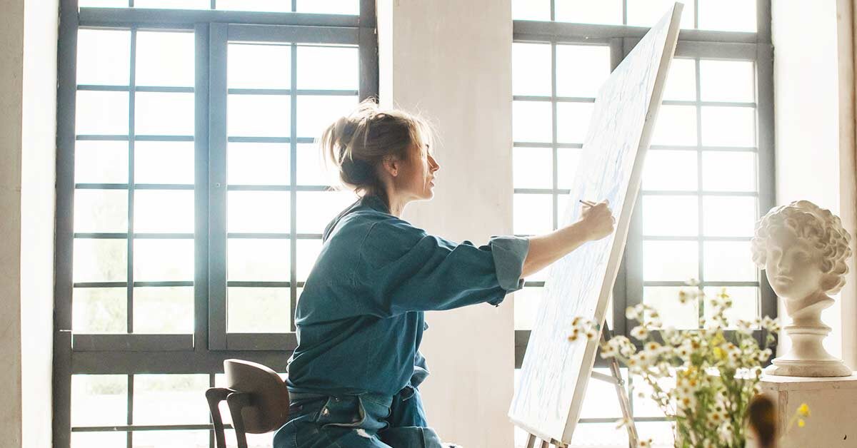 blond woman in blue denim shirt painting in front of sun shining through large windows