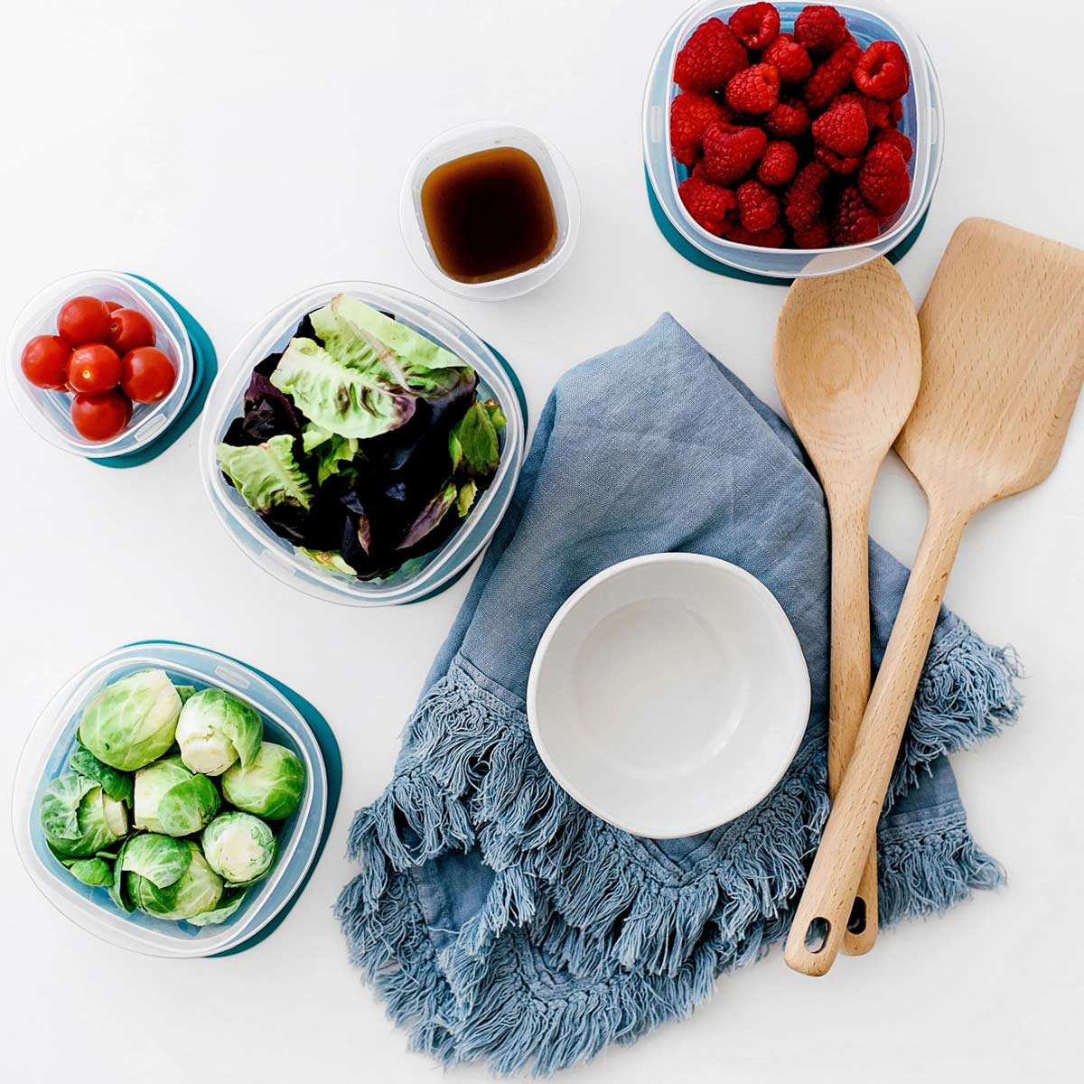 prepared vegetables in containers next to empty bowl and wooden utensils on blue towel sitting on white background