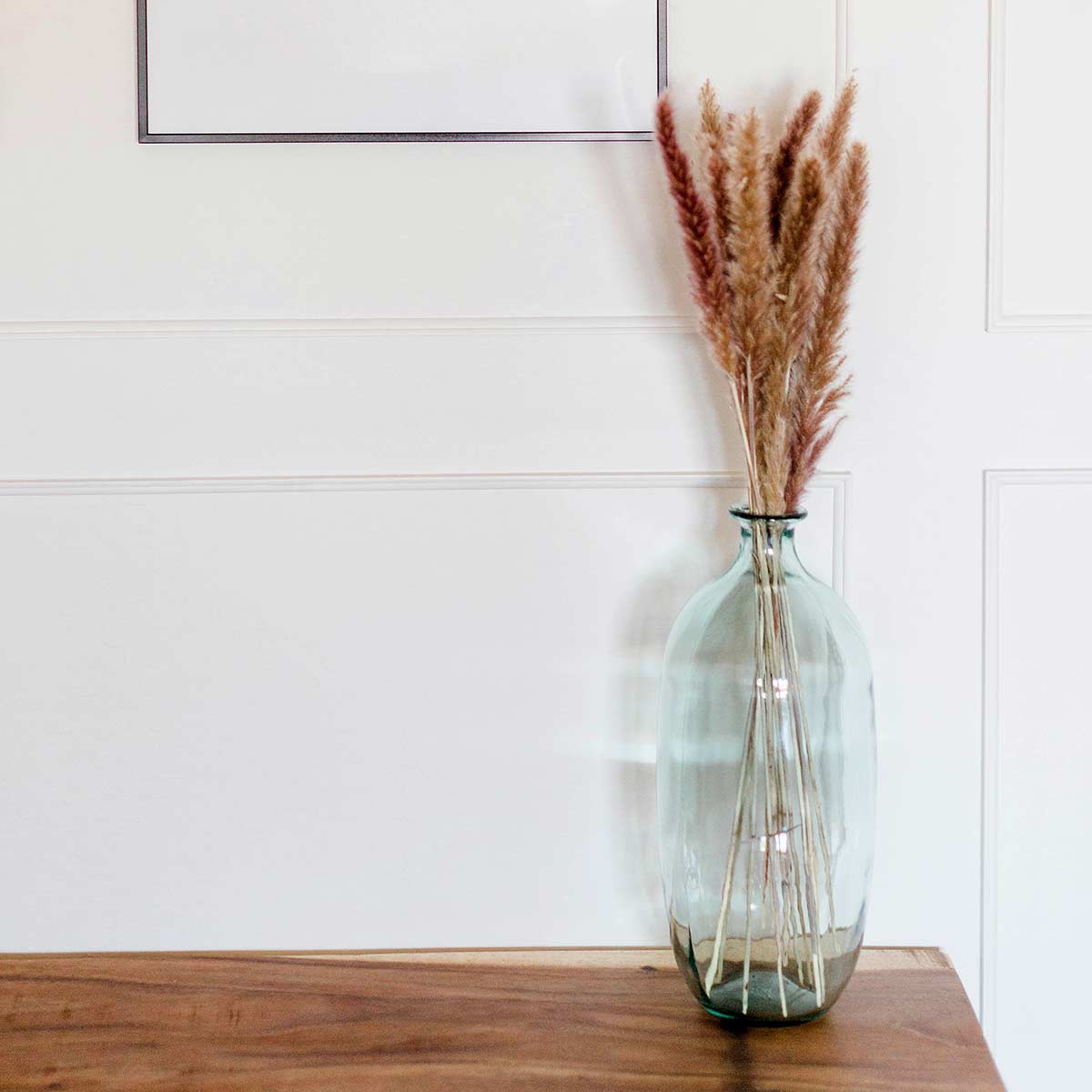 green glass vase with dried grasses on clutter-free wood table against white wall