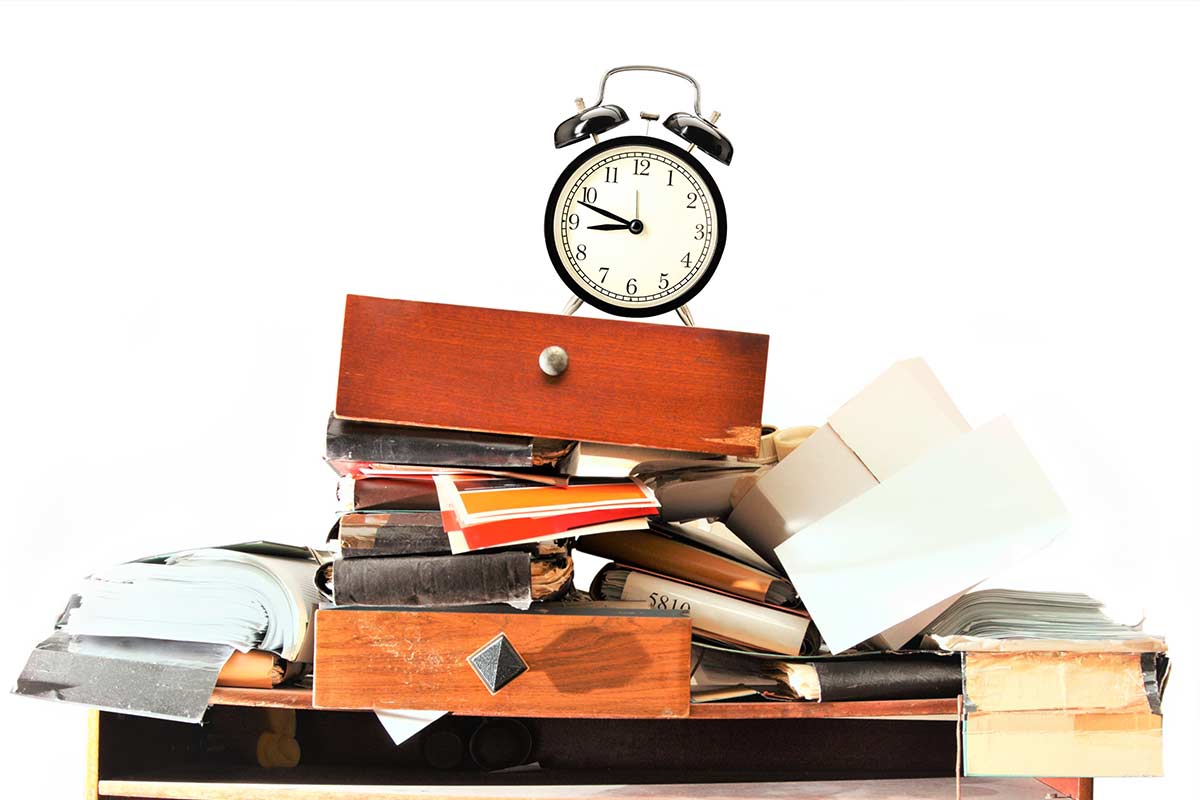 Clock sitting on top of clutter pile of books, papers, and desk drawers against white background.