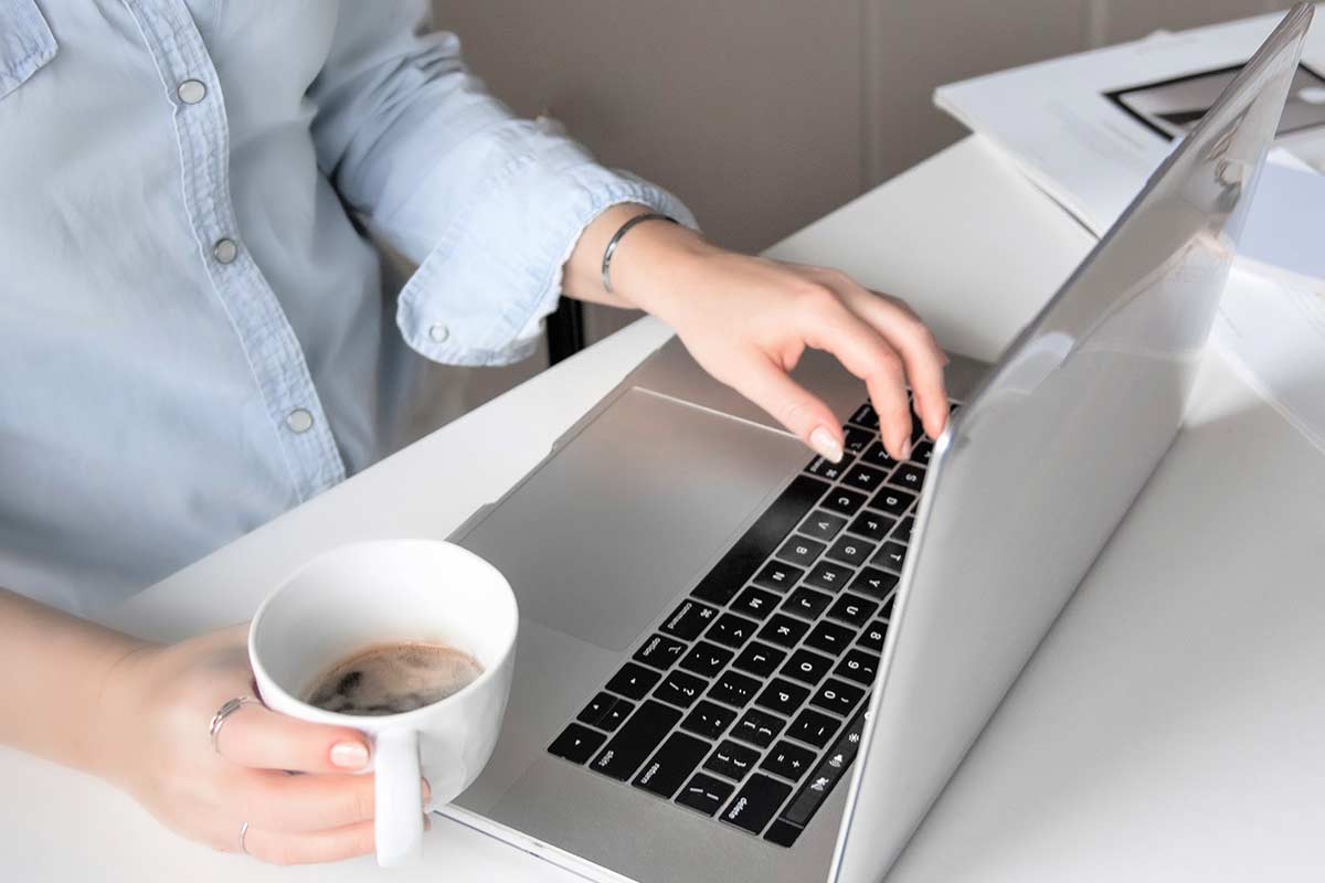 Woman holding white coffee cup and working on laptop computer at white desk.