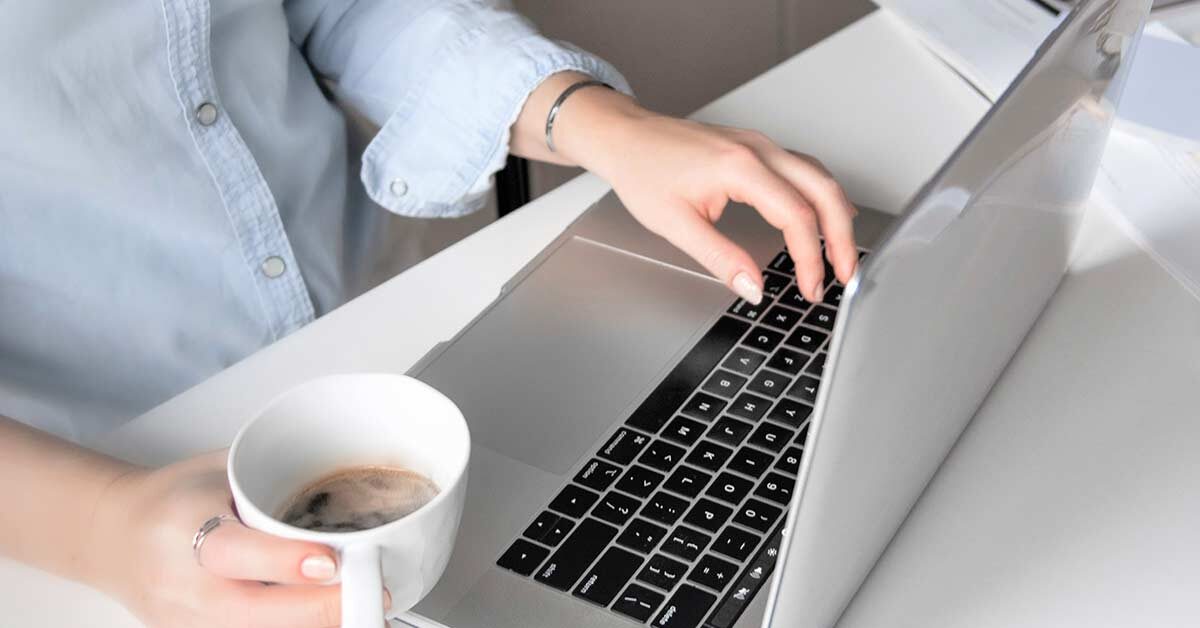 Woman in blue shirt holding white coffee cup and working on laptop computer at white desk.