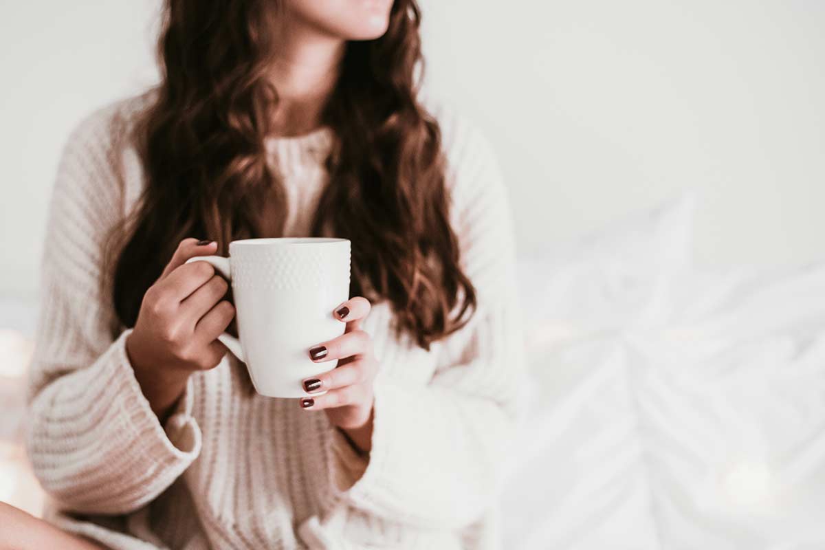 woman with long dark hair wearing white sweater and holding white coffee mug