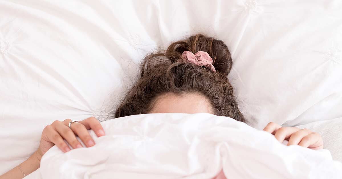 woman with dark hair in pink scrunchie pulling bedcovers over face