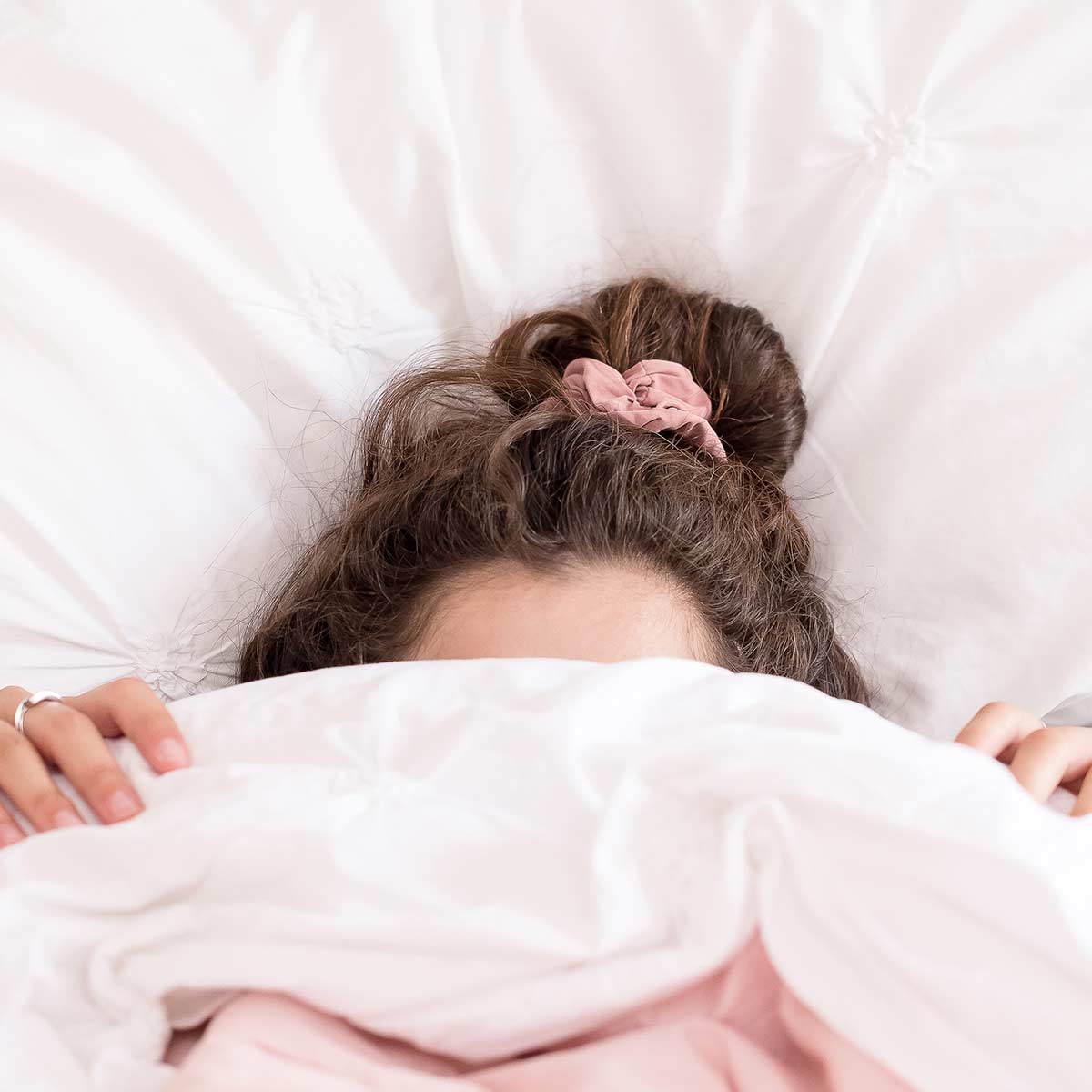 woman with dark hair in pink scrunchie pulling covers over face because she feels too lazy to get out of bed