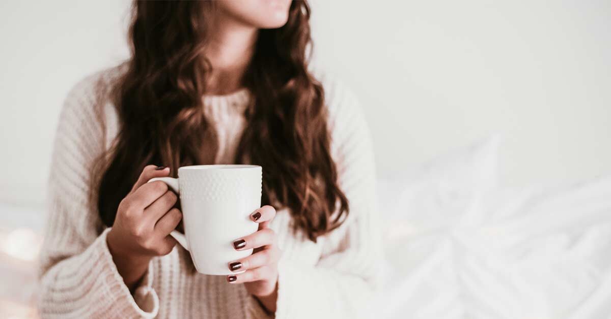 woman with long dark hair wearing white sweater and holding white coffee mug