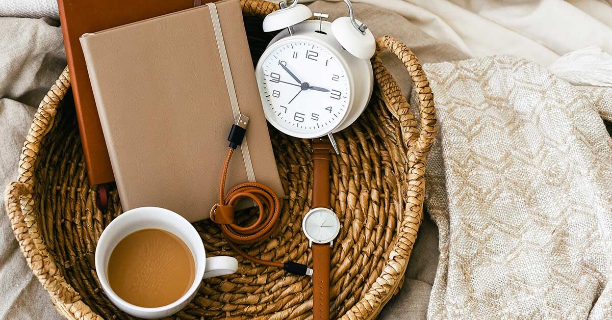 shallow woven basket with watch, coffee, clock, and journals sitting on linens