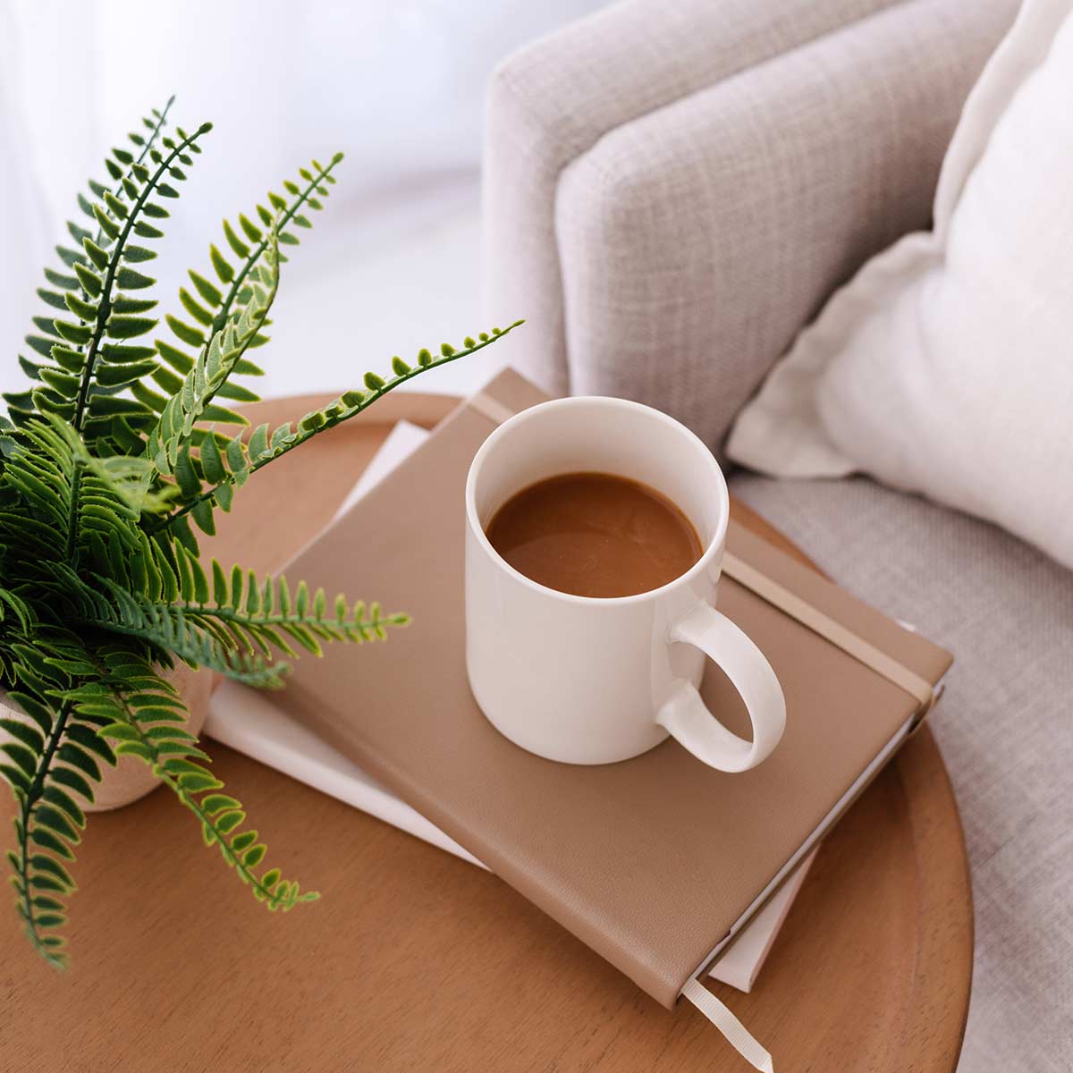 White coffee mug on journal stack sitting on round wood table next to plant in neutral-decor living room.