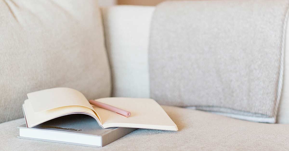 Hardcover book and open journal with pink pen sitting on white fabric cushion with folded woven gray blanket over arm of couch.