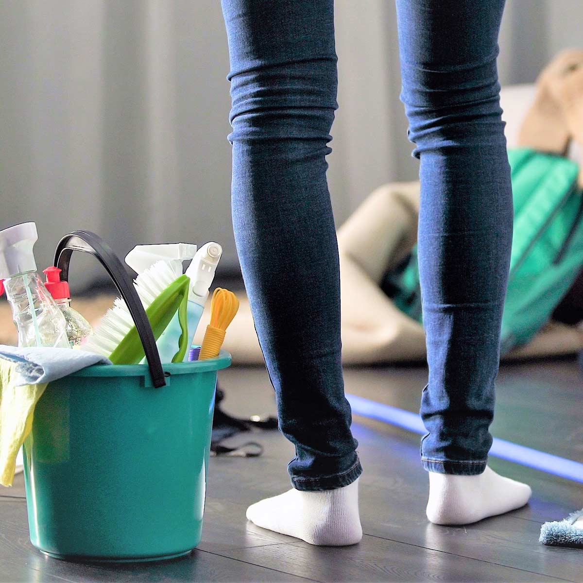 Woman in jeans and white socks standing in cluttered room next to cleaning supplies on wood floor.