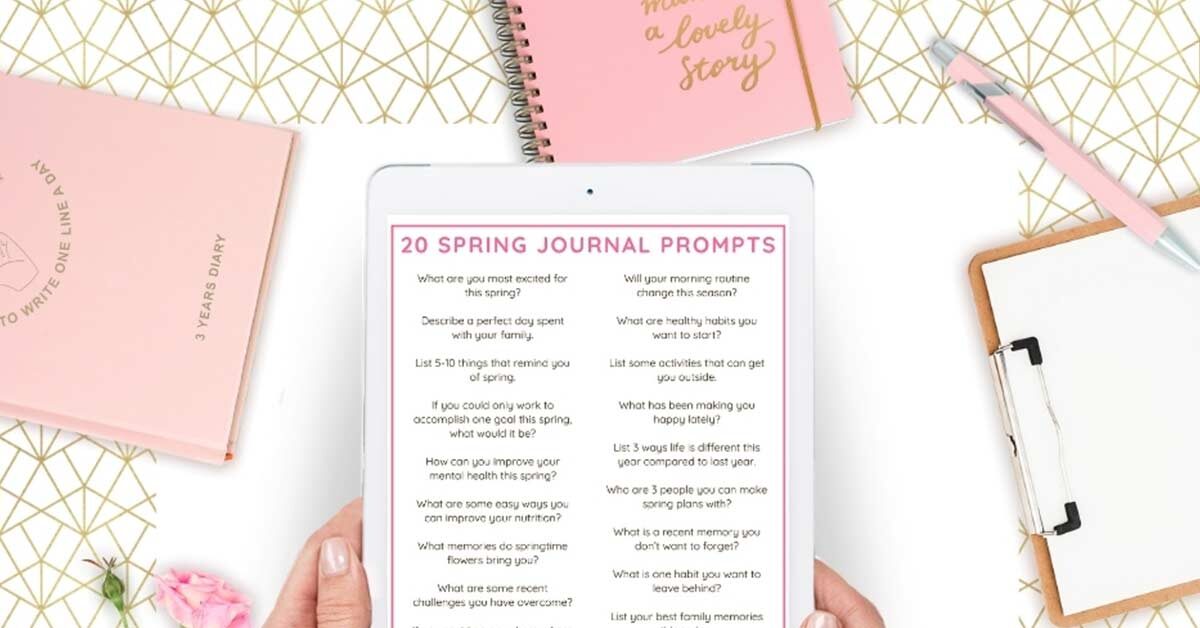 Woman's hands holding digital tablet displaying spring journal prompts above flatlay of pink journals and desk items.