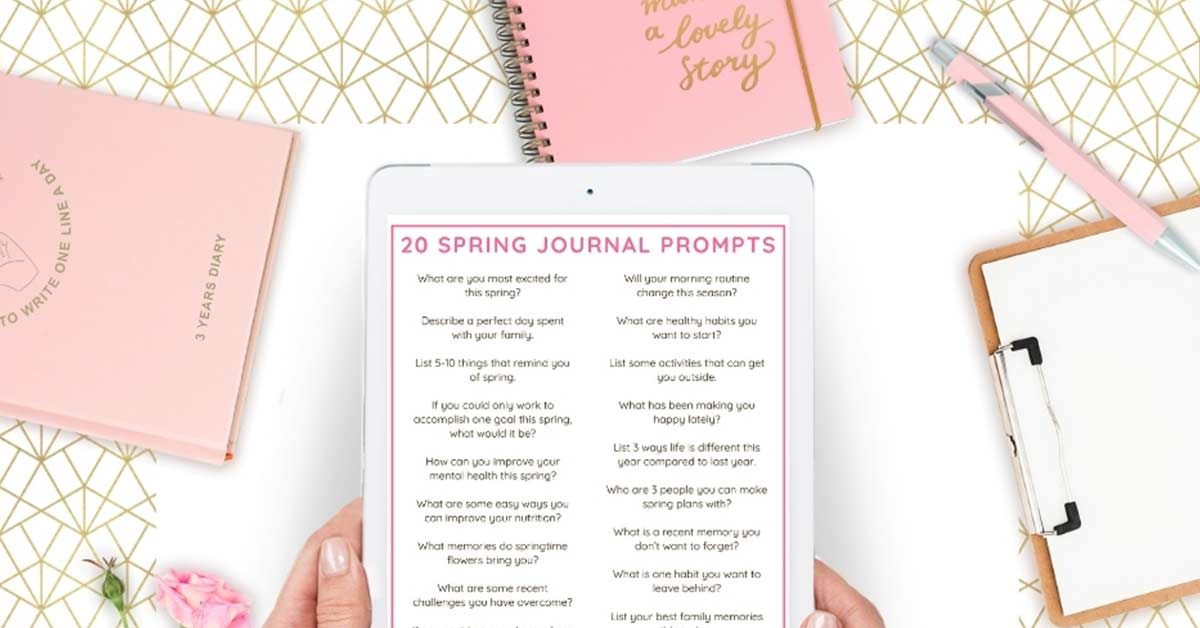 Woman's hands holding digital tablet displaying spring journal prompts above flatlay of pink journals and desk items.