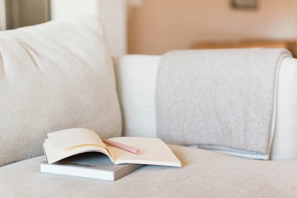 Hardcover book and open journal with pink pen sitting on white fabric cushion with folded woven gray blanket over arm of couch.