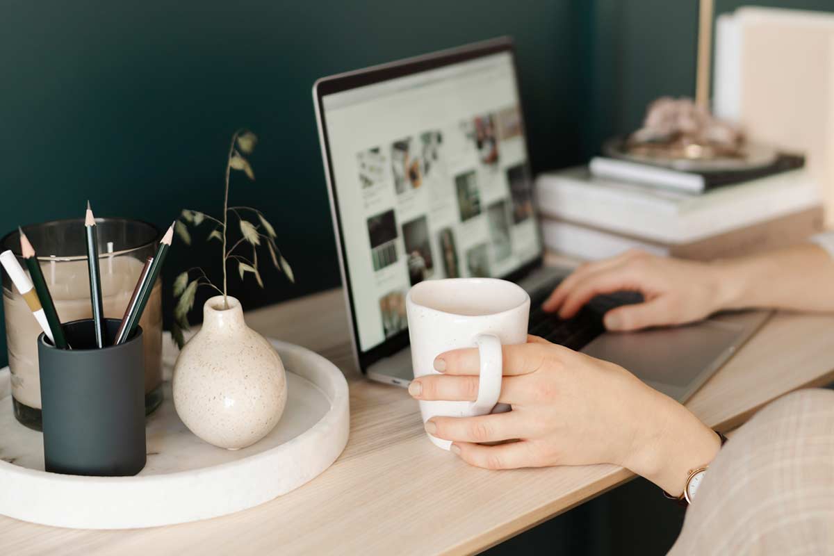 Woman sitting at wood desk and holding coffee mug while scrolling on laptop computer.