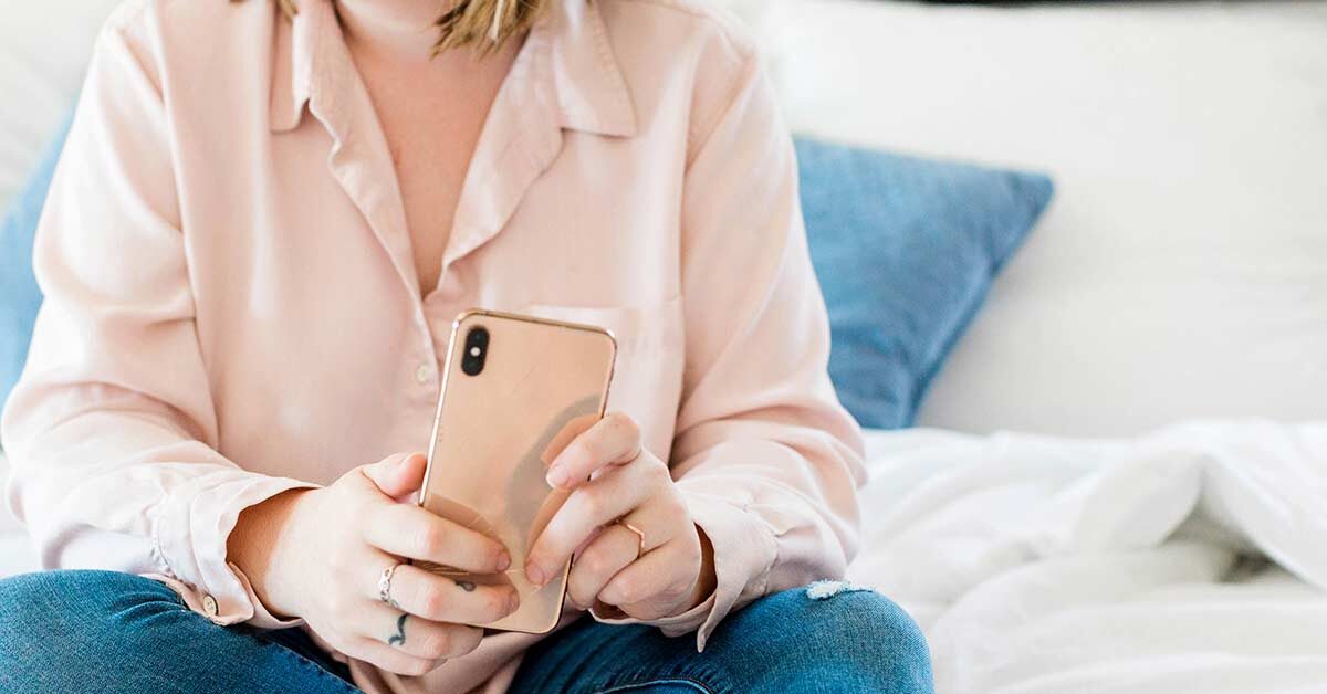 Woman in pink blouse and jeans sitting down while holding pink phone.