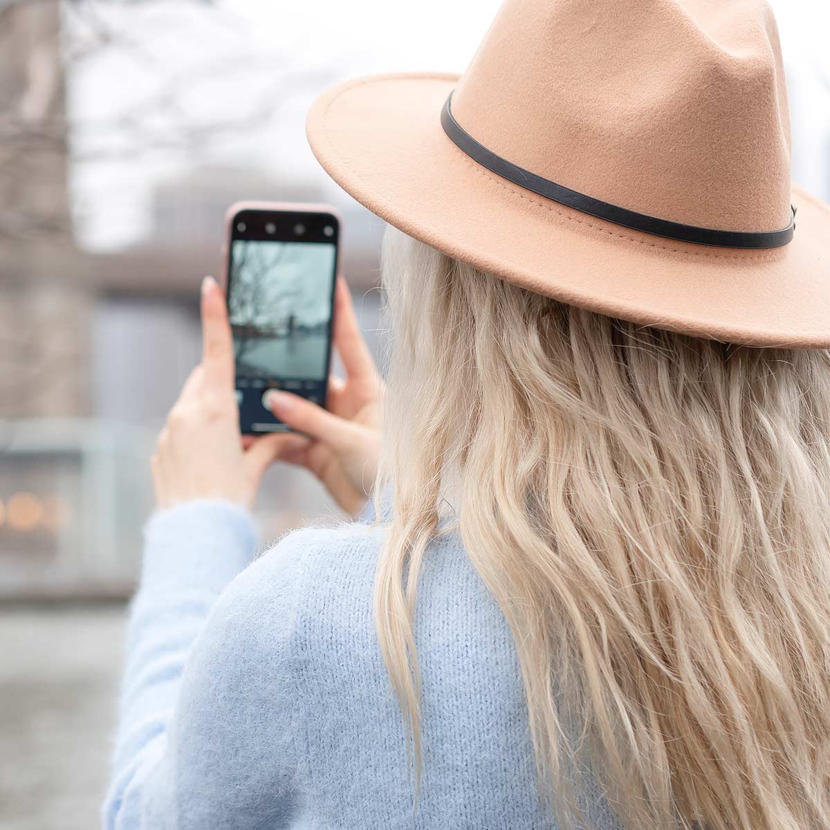 Blonde woman in hat and blue sweater taking outdoor photos on her phone.