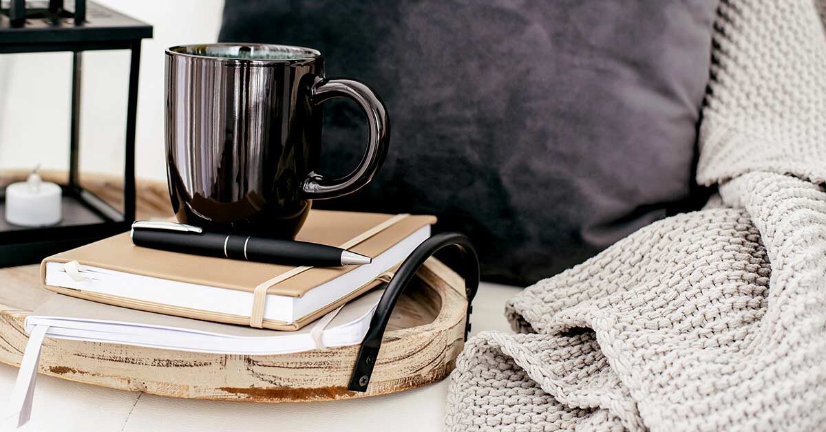 Journals and black coffee mug on wood tray next to knit blanket and throw pillow.