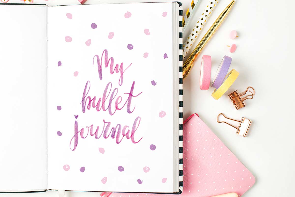 Pink and purple lettered bullet journal page in open journal lying on white desk with pens and other desk supplies.