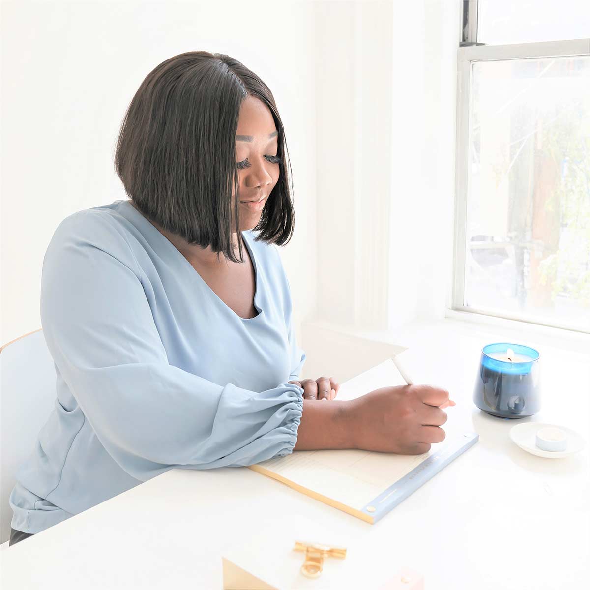 Beautiful Black woman in blue top sitting in bright home office and preparing to do creative journaling.