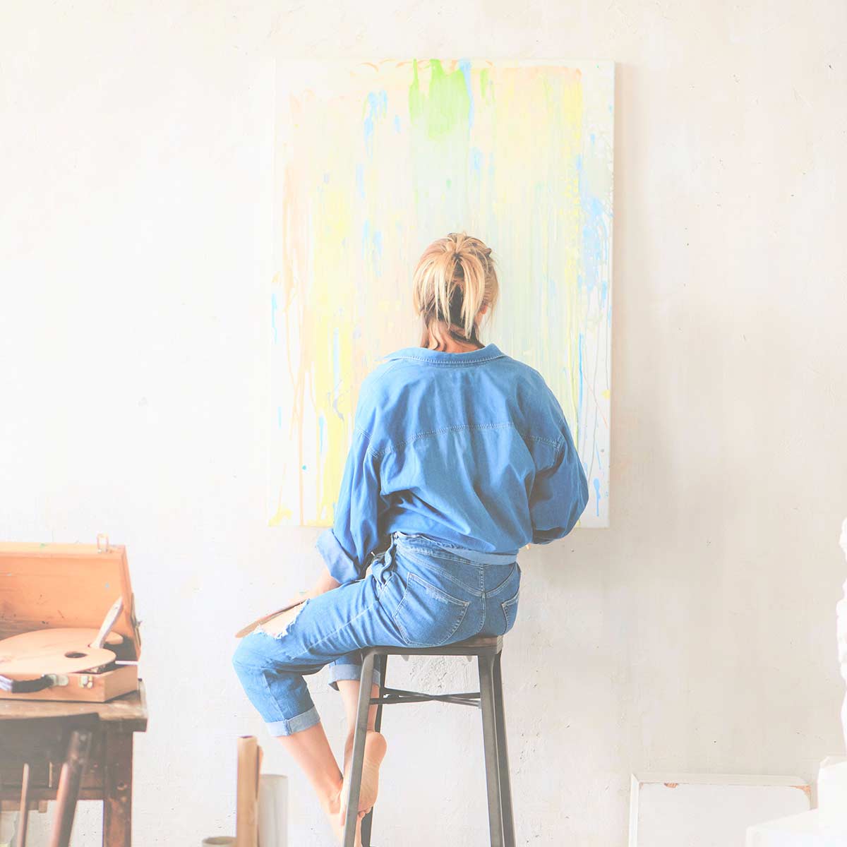 Blonde woman in denim clothing finding creativity by painting a canvas hung on white wall.