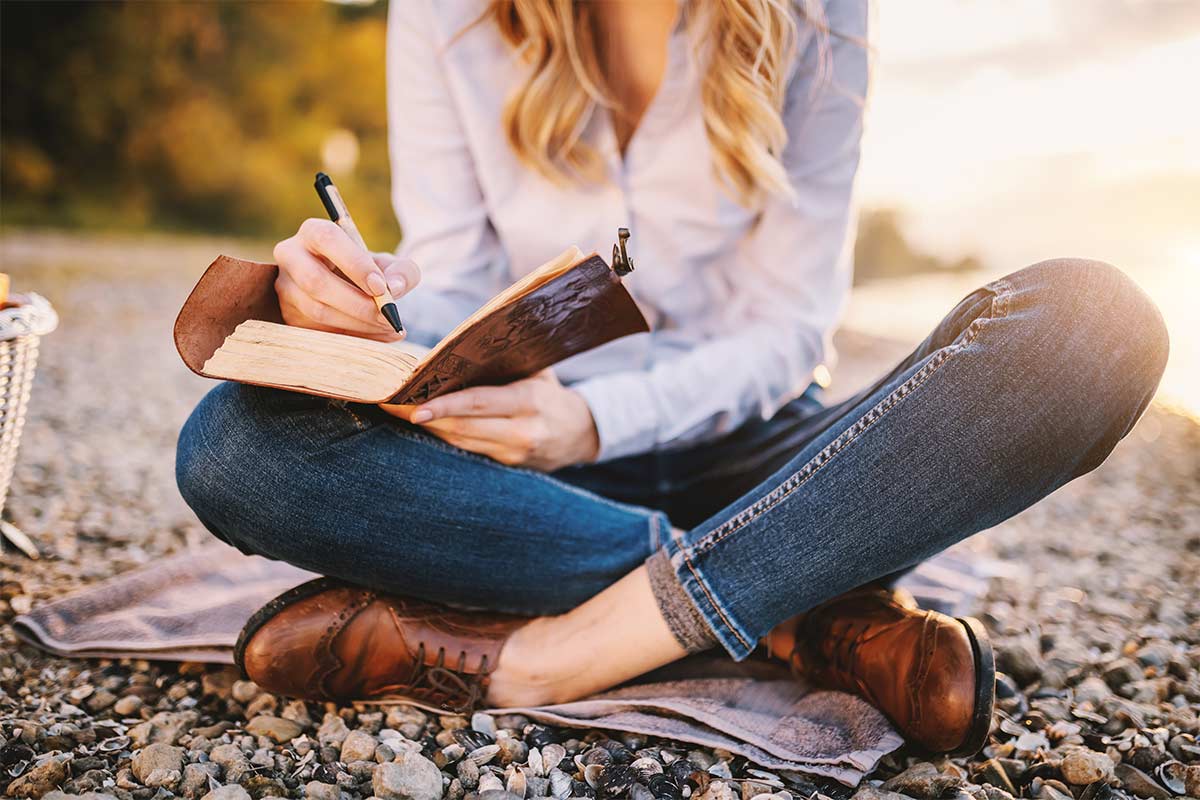 Woman in jeans and blue shirt sitting on ground with legs crossed while writing in leather journal.