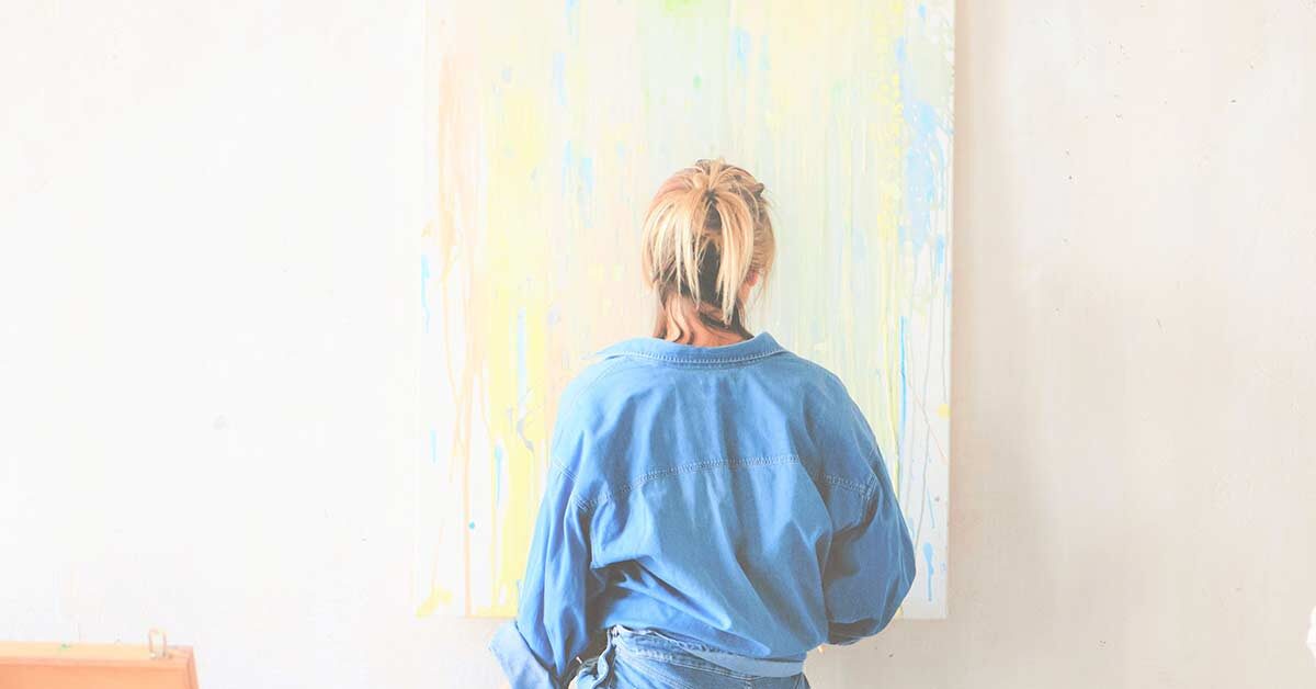 Blonde woman in denim clothing finding creativity by painting a canvas hung on white wall.