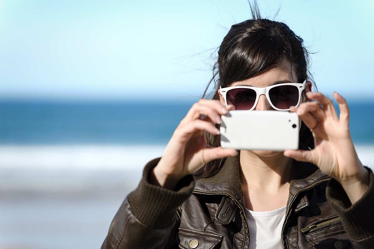 Woman wearing white sunglasses and leather jacket standing outdoors and holding phone up to take a photo.