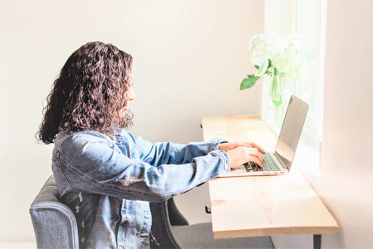 Beautiful Black woman in denim jacket typing on laptop computer in sunny home office.