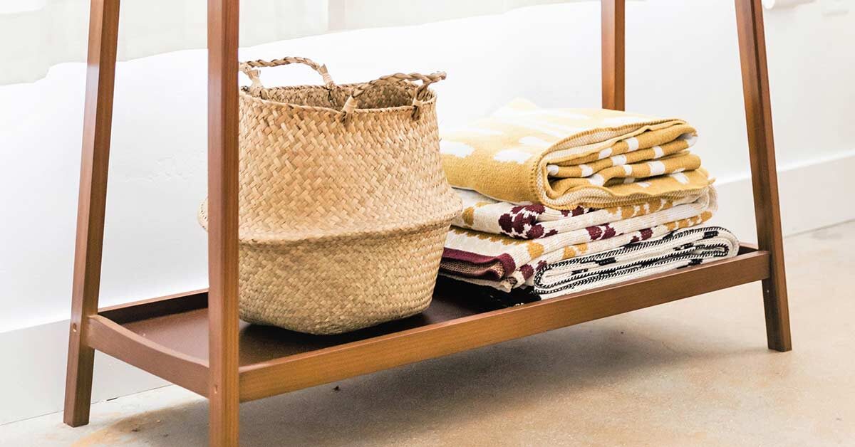 Wood shelf on bottom of table with woven basket and small stack of neatly folded blankets against white wall.