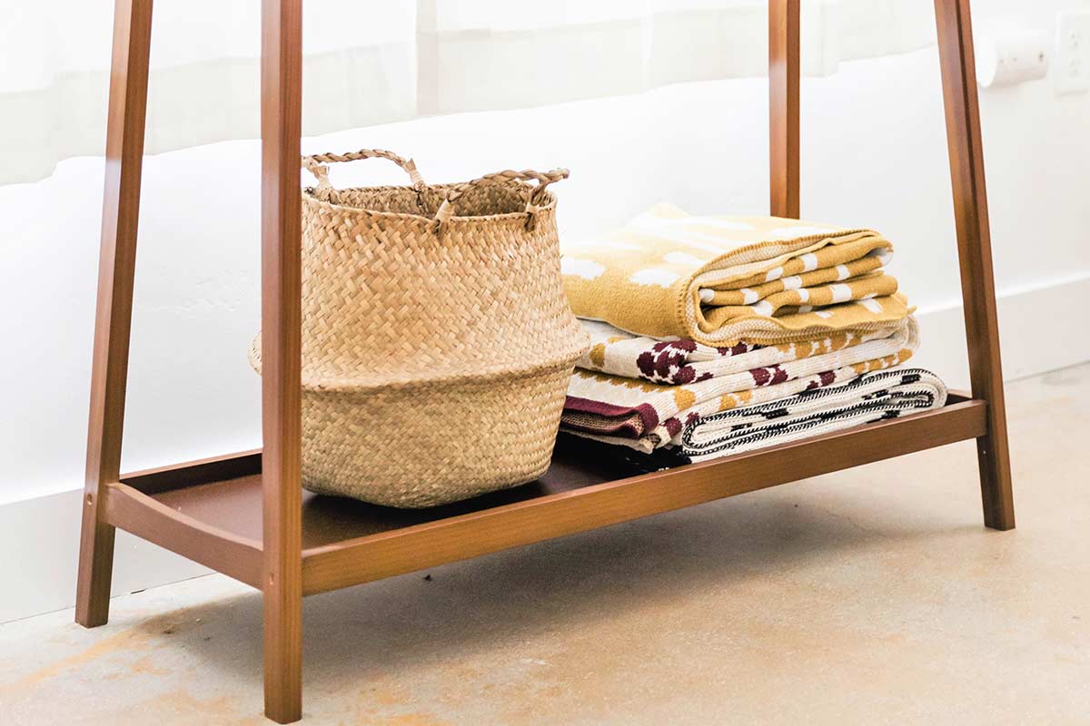 Wood shelf on bottom of table with woven basket and small stack of neatly folded blankets against white wall.