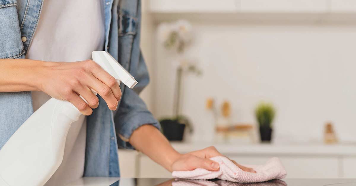 Woman in blue chambray shirt and white tee holding spray bottle and wiping glass stovetop with pink cleaning cloth.
