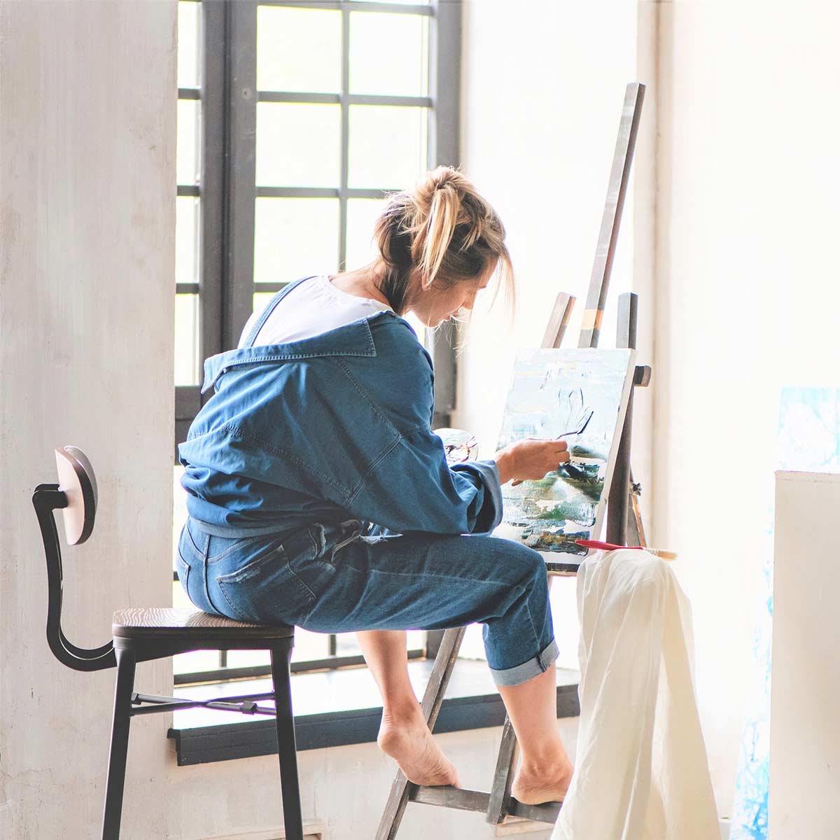 Blonde woman in denim clothing painting in sunny home studio.