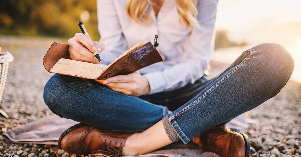 Woman in jeans and blue shirt sitting on ground with legs crossed while writing in leather journal.