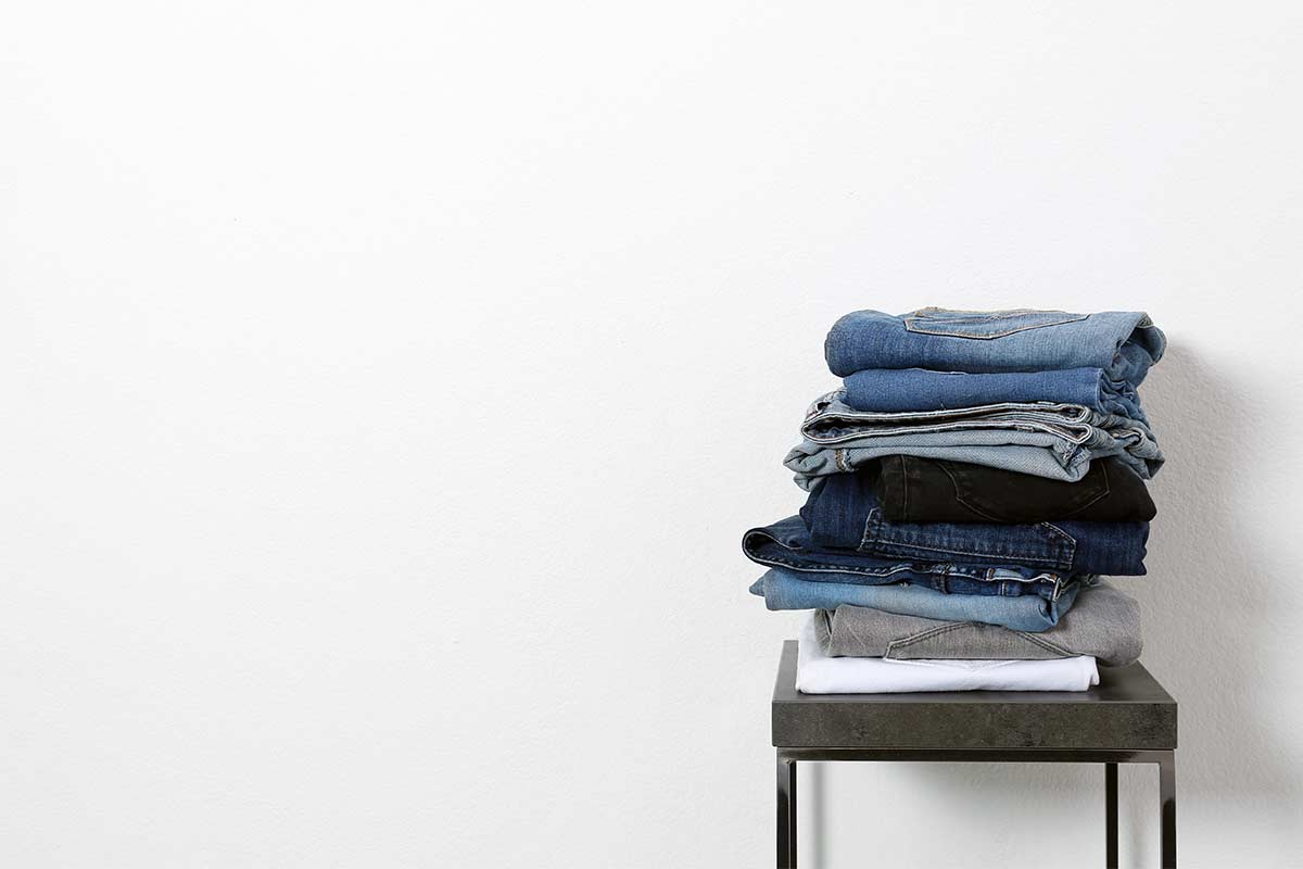 Pile of folded jeans on stool against white wall.