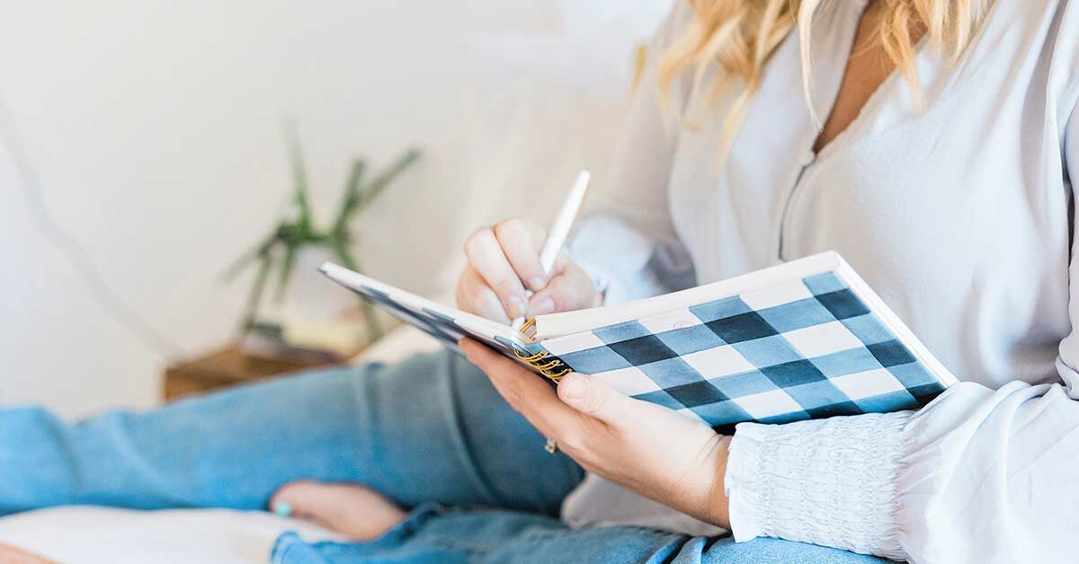 Woman with long blond hair wearing blue blouse and denim jeans sitting and writing in black and white buffalo check journal.