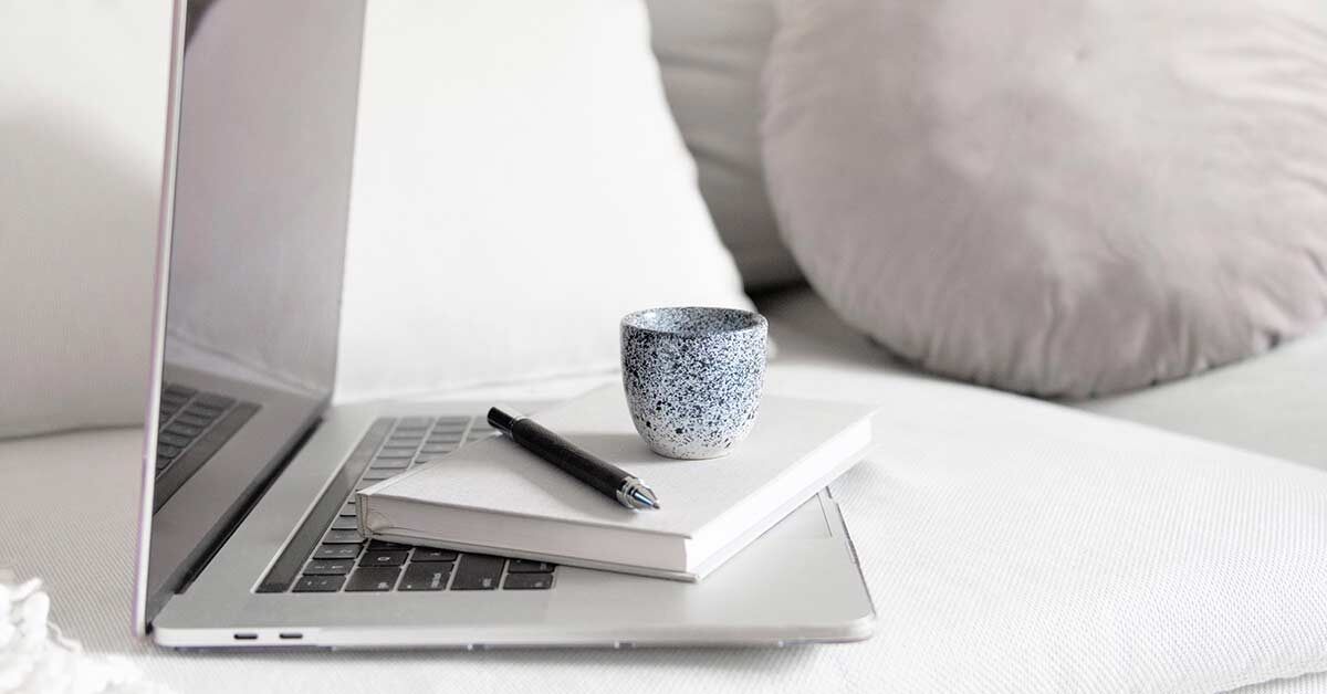 Journal, pen, coffee mug stacked on open laptop sitting on white couch.