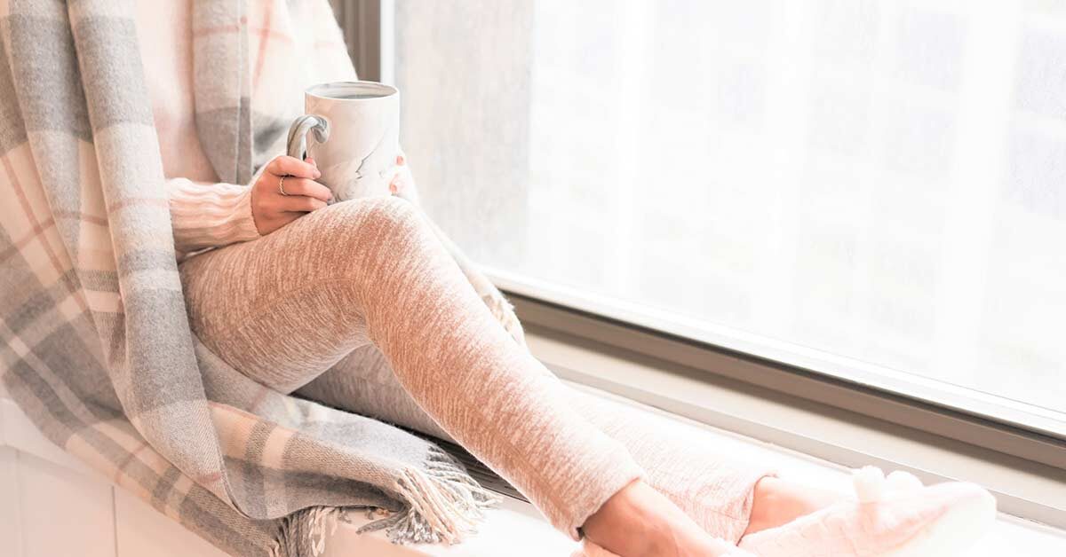 Woman wearing cozy clothes, slippers, and blanket while holding coffee mug and relaxing next to window during winter break.