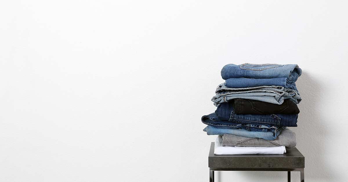 Pile of folded jeans on stool against white wall.