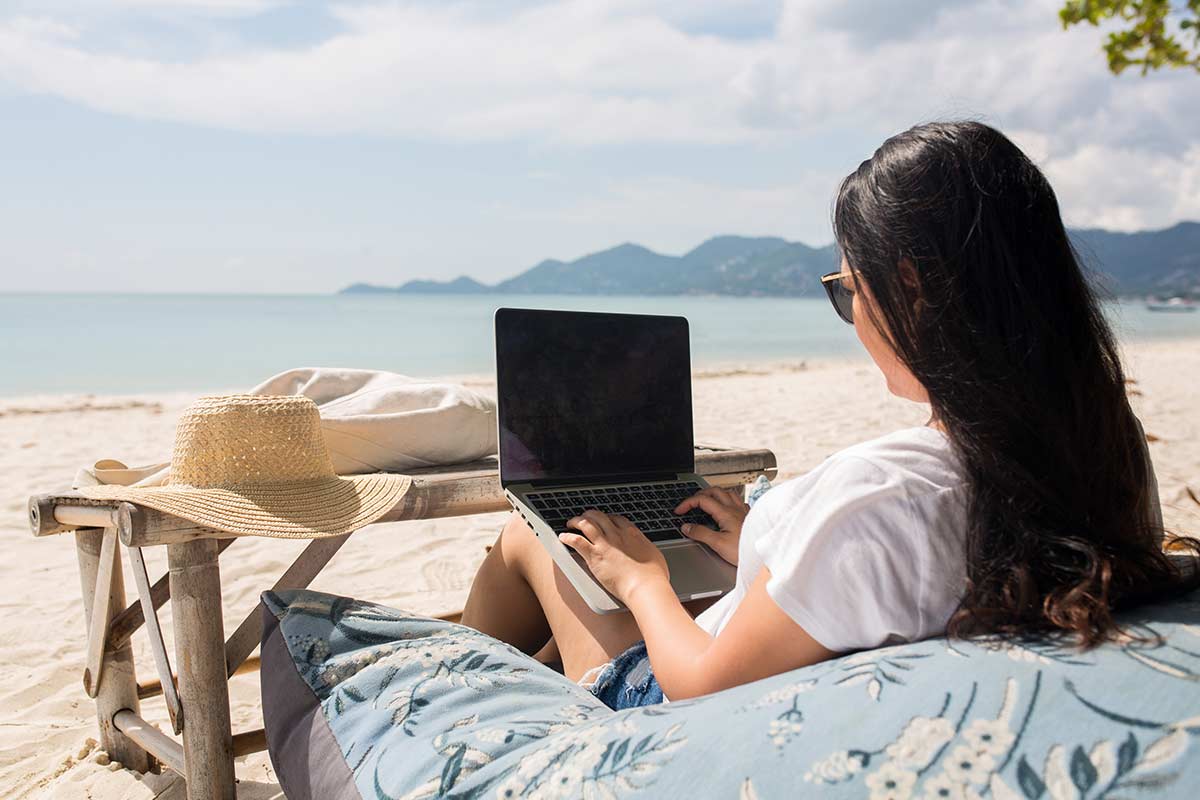 Woman with long black hair and white tee shirt sitting on huge pillow while being productive on her laptop at the beach.