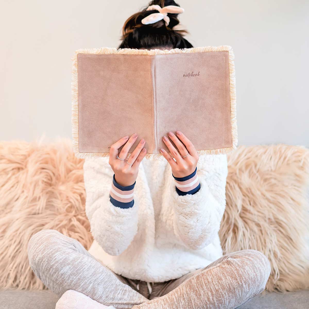 Woman sitting cross-legged on couch in front of fuzzy pillows and holding up old journal to read over winter break.