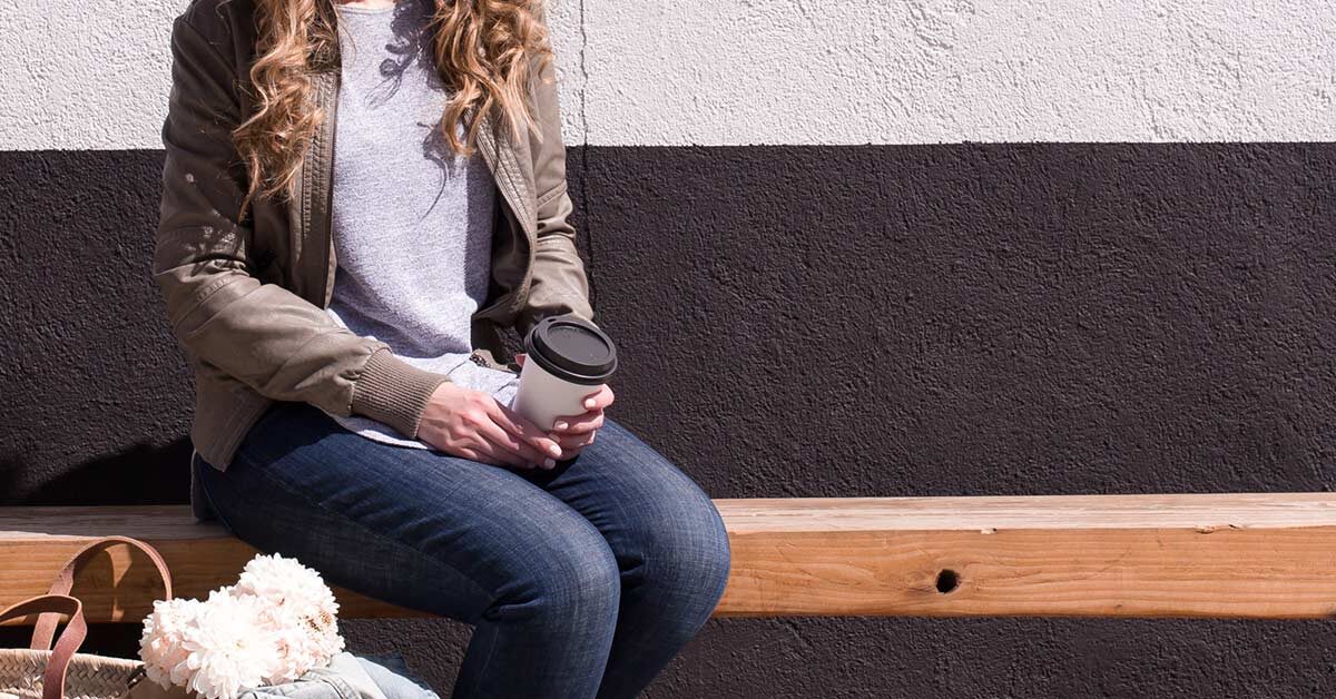 Woman in casual clothing sitting on wood bench outdoors while holding coffee cup and taking a break.